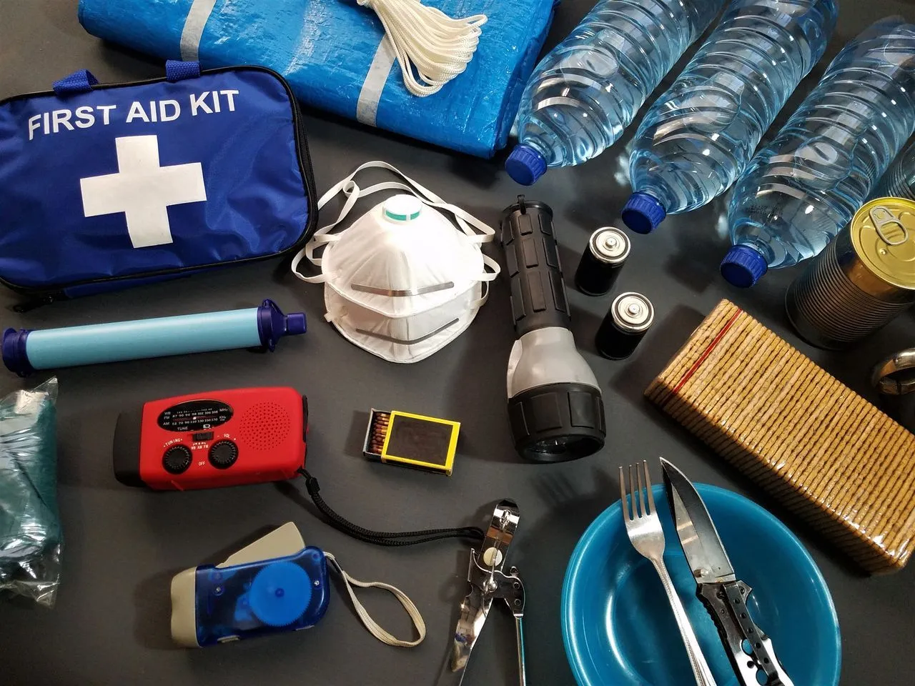 Emergency kit supplies laid out flat: first aid kit, bottled water, hand-crank radio, flashlight, batteries, N95 masks, and a manual can opener