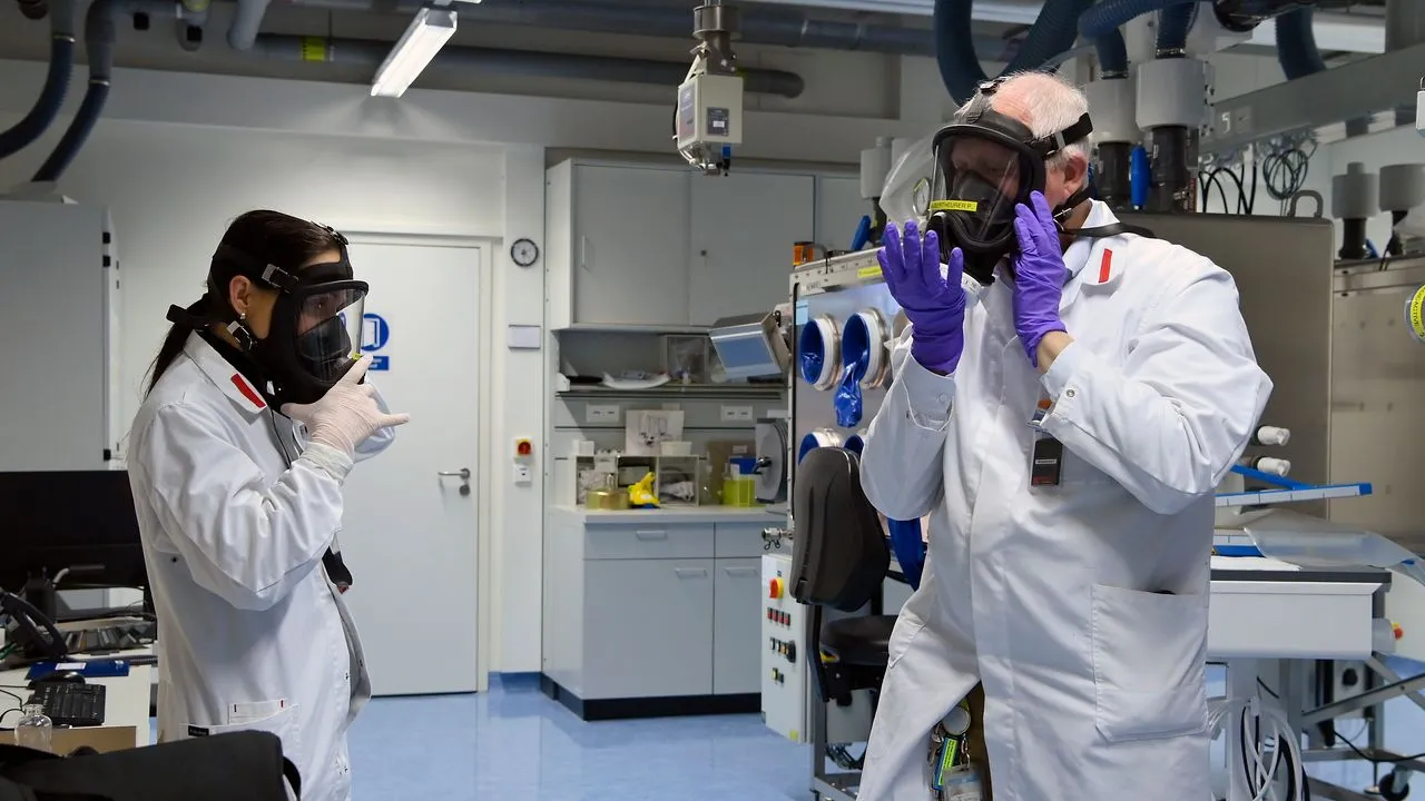 Two laboratory technicians in white coats putting on full-face respirators in a bright industrial lab