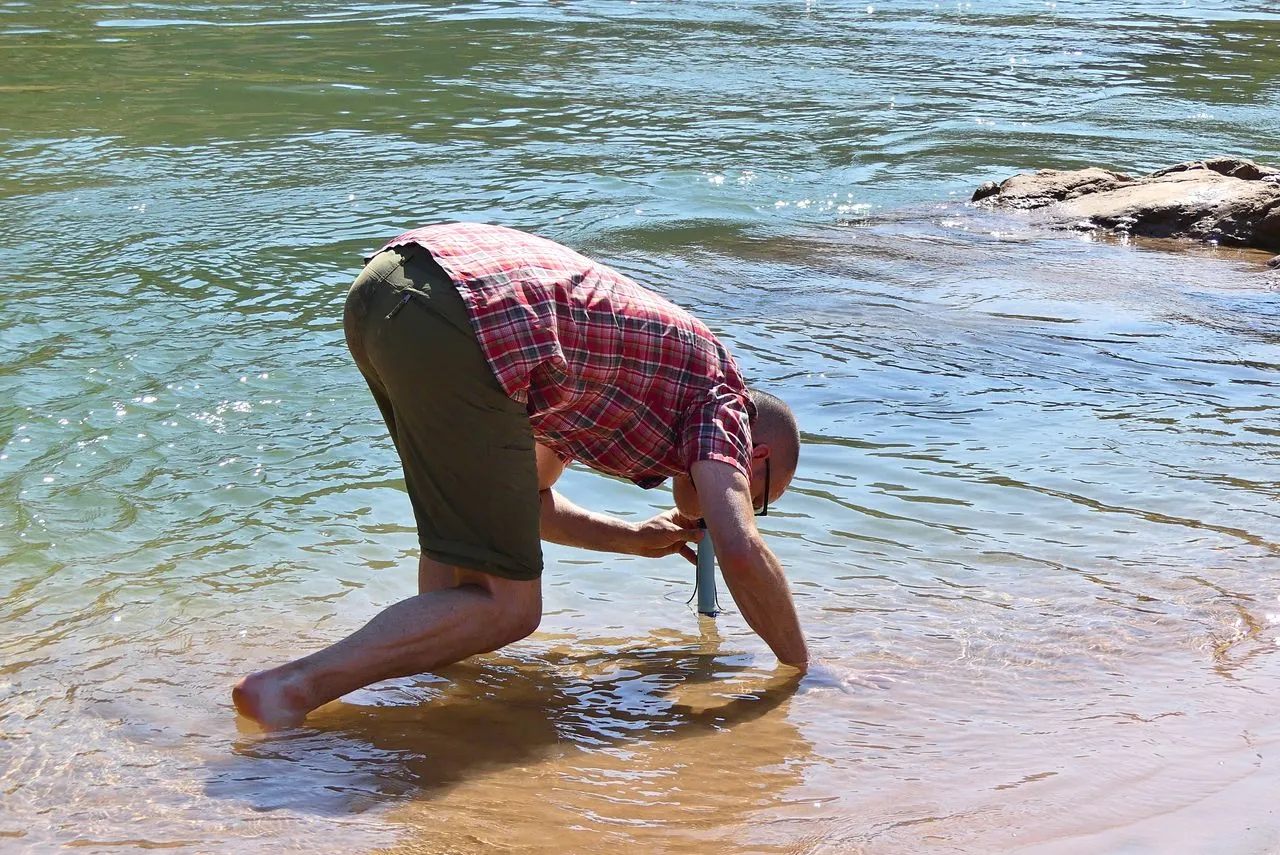 Hiker kneeling at a river's edge drinking through a personal straw water filter