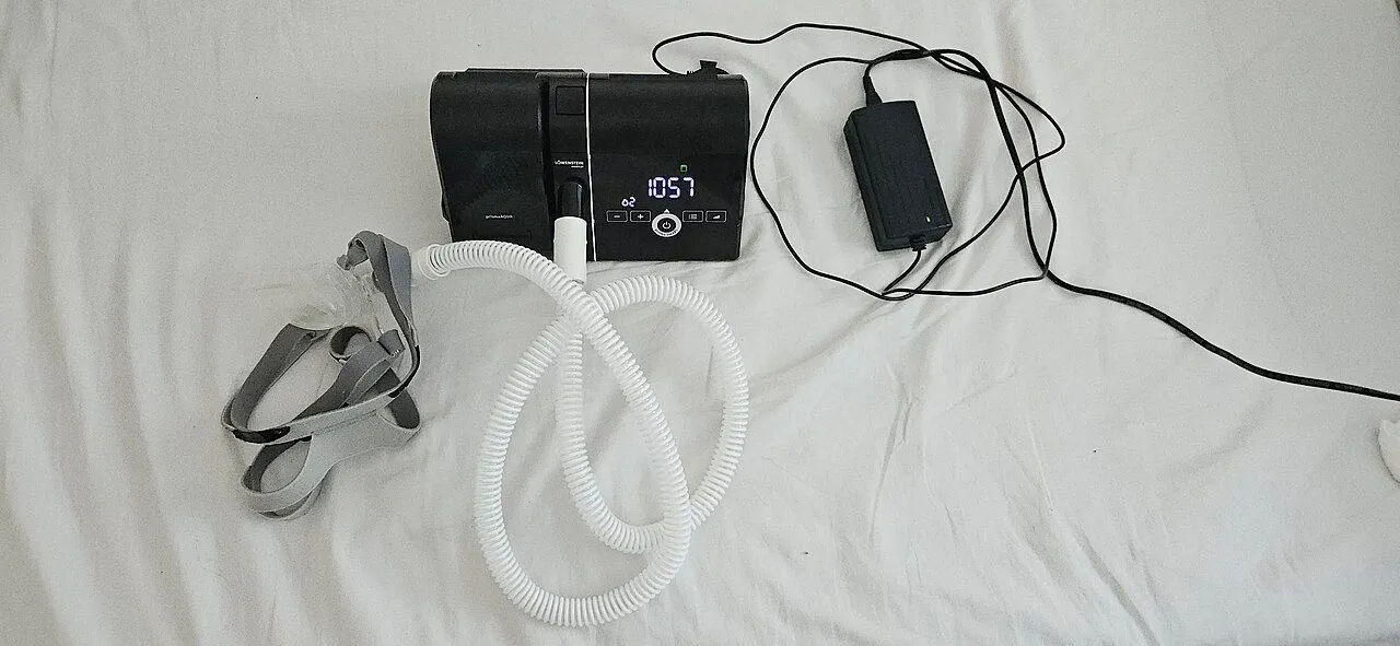 CPAP machine with hose, headgear and its power adapter laid out on a white bed