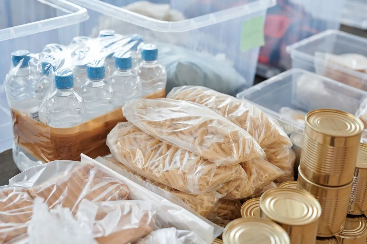 Emergency food stockpile with bottled water, bagged pasta, canned goods, and clear storage bins